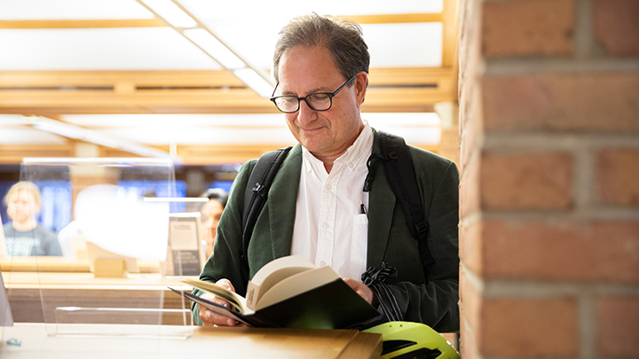 Middle-aged man with dark glasses, white shirt, green blazer, and neon green biking helmet stands at end of library shelf with an open book with green cover.