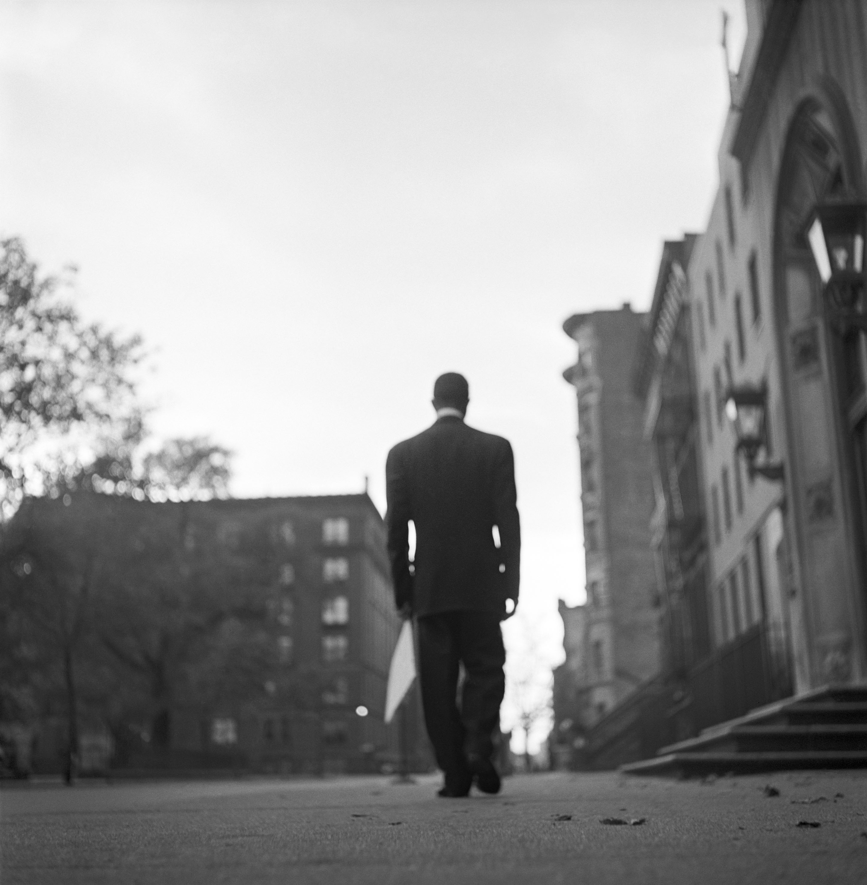 Slightly unfocused image of a man in suit walking through a setting of apartment buildings and trees. Man is seen from behind partly silhouetted against a white sky.