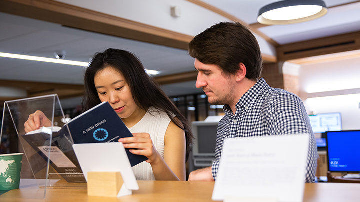 Woman with long dark hair and white sleeveless top stands next to man with brown hair and slight beard wearing blue and white check shirt, as both look at a book title "A New Dawn"