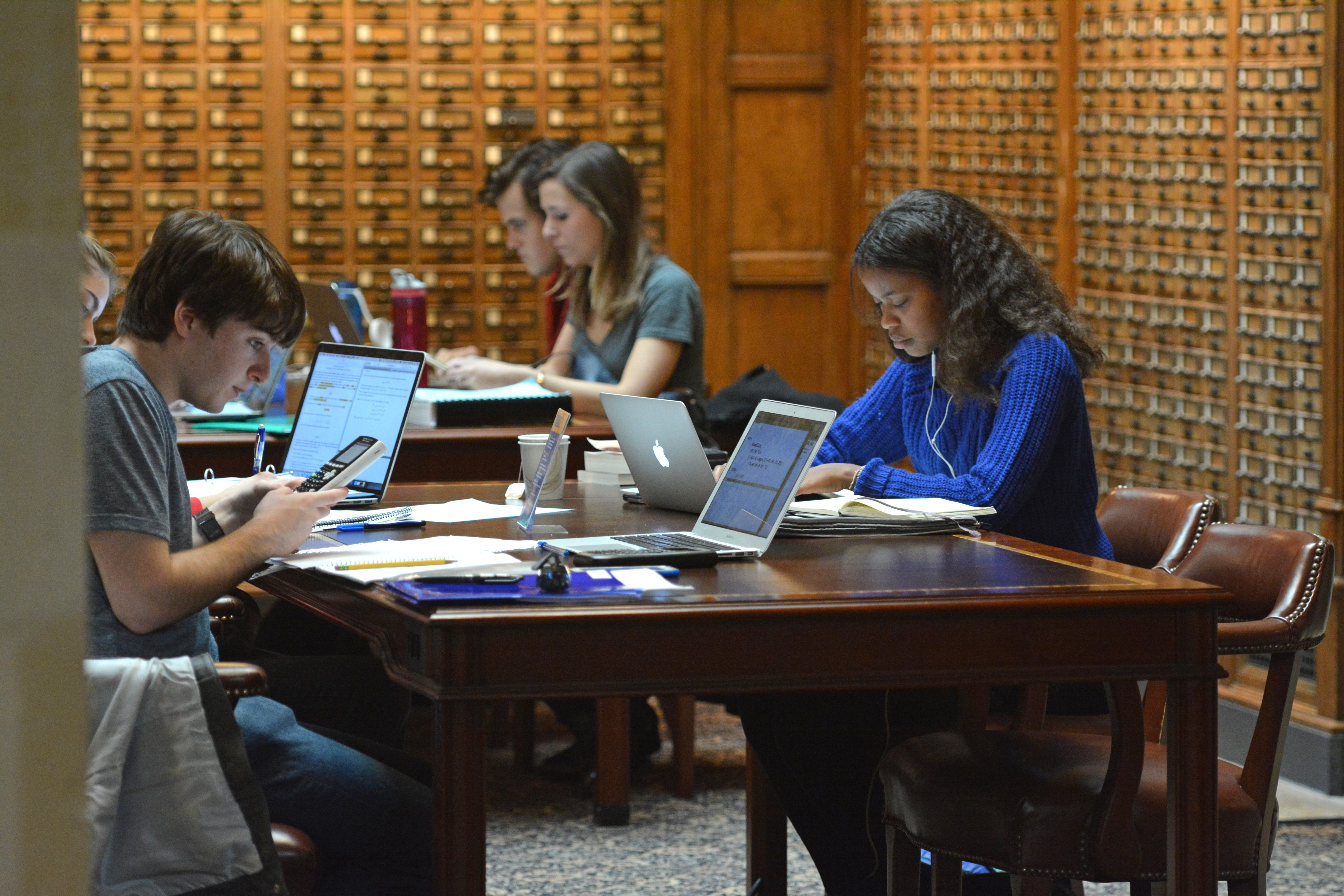 Students with laptops studying at library tables, background of card catalogs
