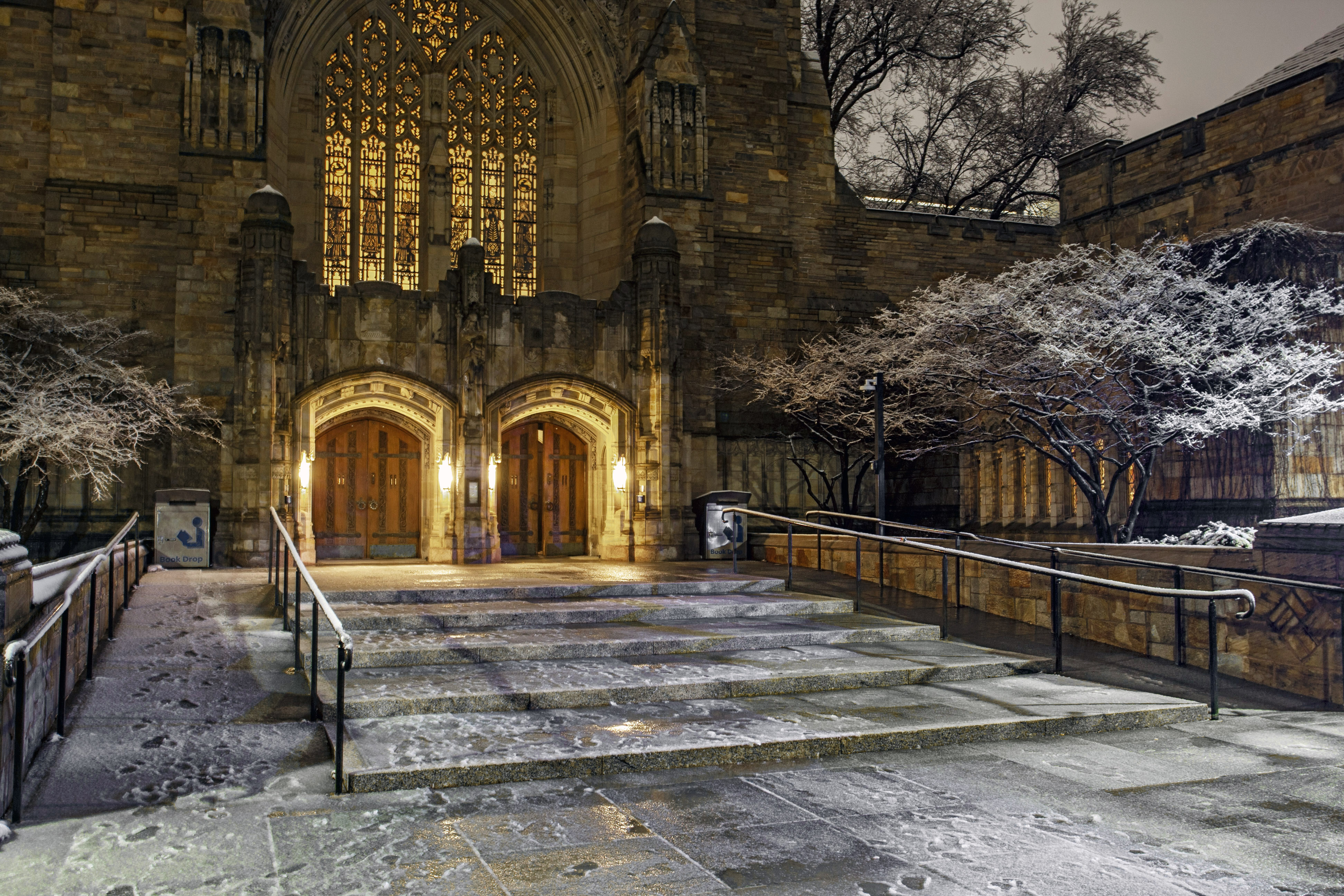 nighttime scene of library entrance with light over arched doors and through stained glass windows and light snow on the ground