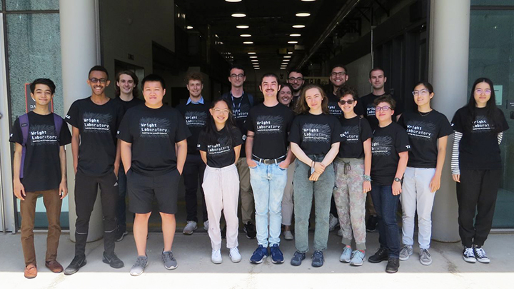 17 young male and female science students pose, wearing black t-shirts with white lettering that reads "Wright Laboratory"