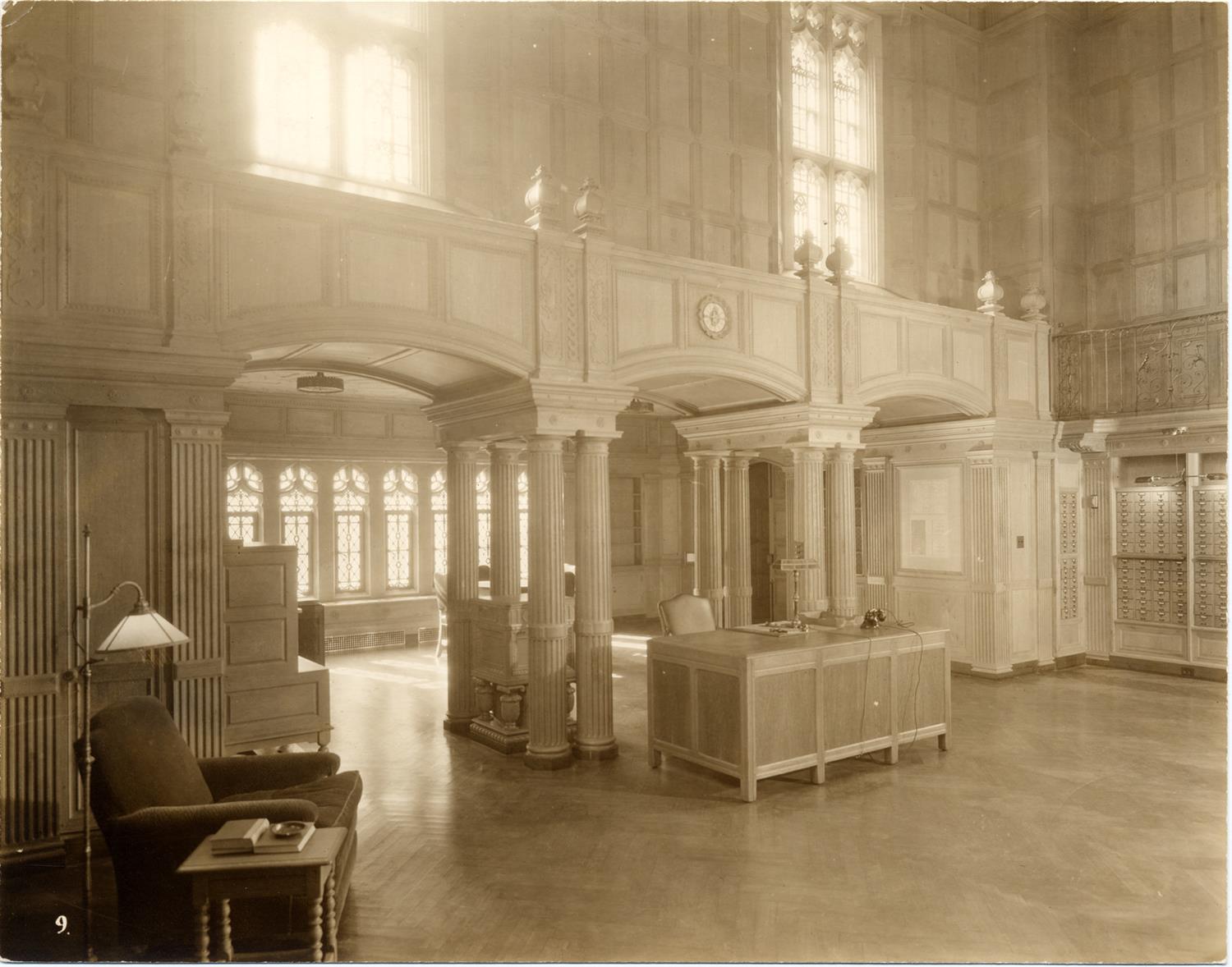 Monochrome image of a room with arched entrance, librarian desk and other old-fashioned furnishings.