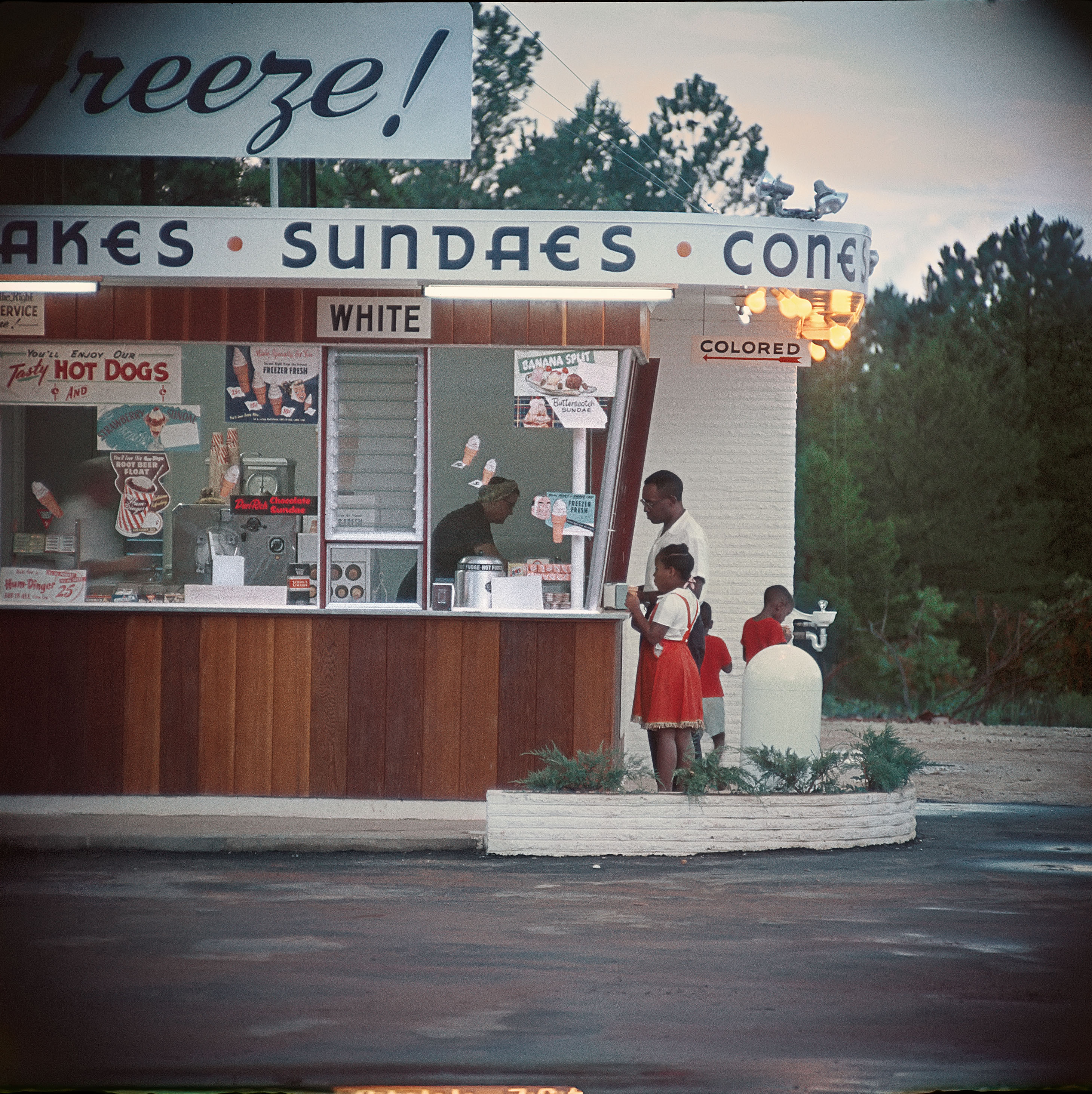 Black man and children being  served at an ice cream stand under a sign reading "colored". A sign above another window reads "white" .