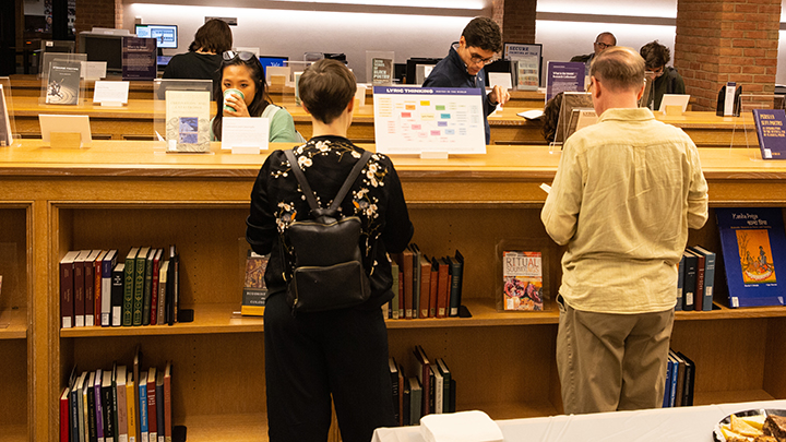 Seven people peruse three rows of books on display. The two people closest to camera have their backs to the viewer. The woman wears a black embroidered jacket and black leather backpack. She has short brown hair. The man to her right has a yellow long-sleeve shirt and brown pants and has brown hair.