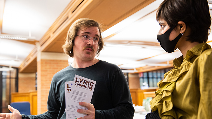Man with jaw-length brown hair, mustache, and wire-rimmed glasses talks to woman wearing ruffled gold blouse, black face mask, and small gold earrings. He holds cards that read "Lyric Thinking"
