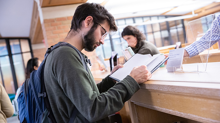Young man learns over book that rests on top of library shelf. He has green sweatshirt and blue backpack, dark hair and beard and dark glasses. In background another man is seen resting on the library shelve reading signage.