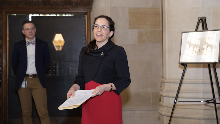 Woman with dark hair and glasses, black blouse, red skirt, holding papers and speaking with stone walls and pillar in background.