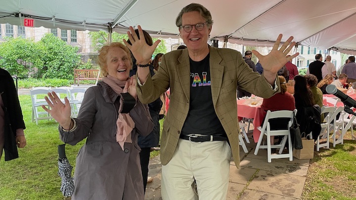 Woman in gray coat and pink scarf and man in brown blazer smiling and posing with uplifted "jazz hands." They are under a tent and in the background are people gathered eating at tables with pink tablecloths and white folding chairs. 