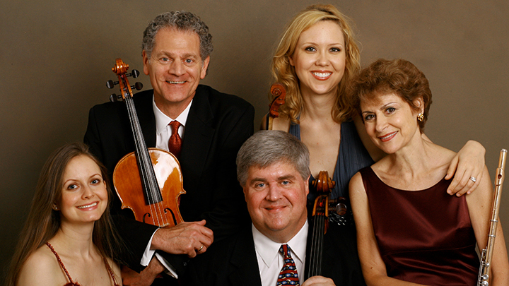Five musicians, three women and two men, pose for camera. One man holds viola, woman with short red hair holds flute