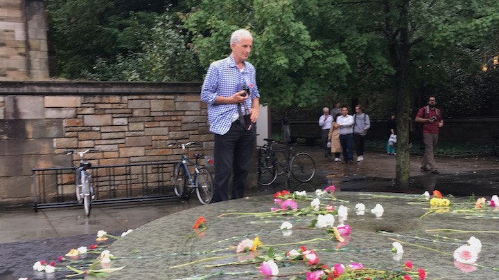 Thin white-haired man in blue checked shirt and dark pants holds camera and looks at stone fountain strewn with flowers.