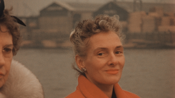 Woman wearing orange coat with salt and pepper brown curls on top of her head looks at camera smiling. She also wears orange lipstick. Behind her is a body of water and a row of blurred buildings. Next to her is a partial view of a woman with short dark hair, wire glasses, wearing fur collar. Only her left side of face is visible.