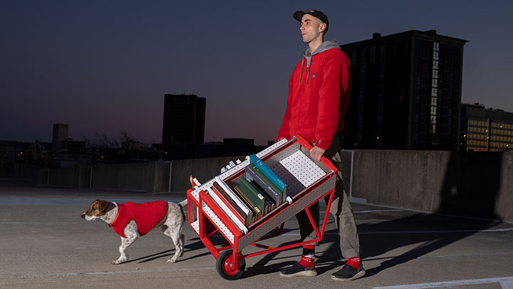 Tall man with red hoodie walking across dark skyline, wheeling a red cart with shelves and walking a brown and white dog wearing a red coat