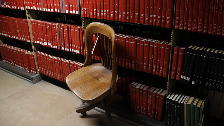 Wooden library chair in front of row of red books with one book tipped between slat of chair back
