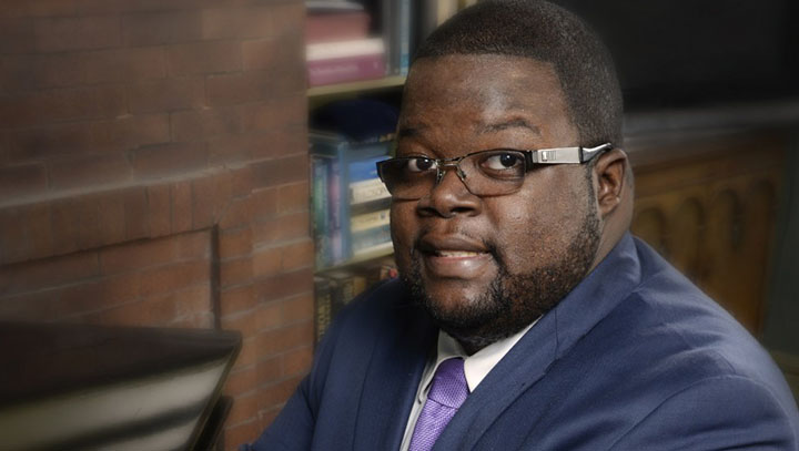 Black man with close cropped beard and hair wearing brown and metal glasses looks into camera. He wears a blue jacket, white shirt, and purple tie.
