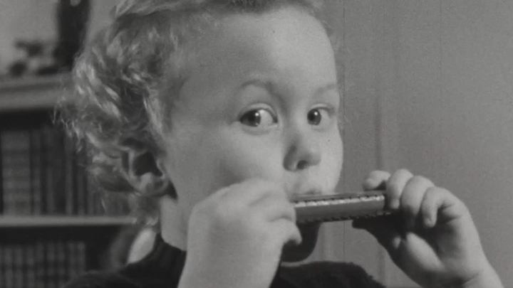 Black and white photo of little girl with curly hair, wide-eyed, blowing into a harmonica that she holds with both hands