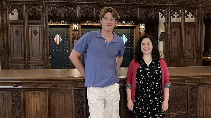 Tall young man with wavy brown hair, wearing wire-rimmed glasses, a blue shirt, white pants, and short necklace, stands next to small woman with dark hair, black and white flowered dress and red sweater. In the background are ornate wood-carved panels and they lean against a long, dark wood-carved surface that had once been the library's circulation desk..