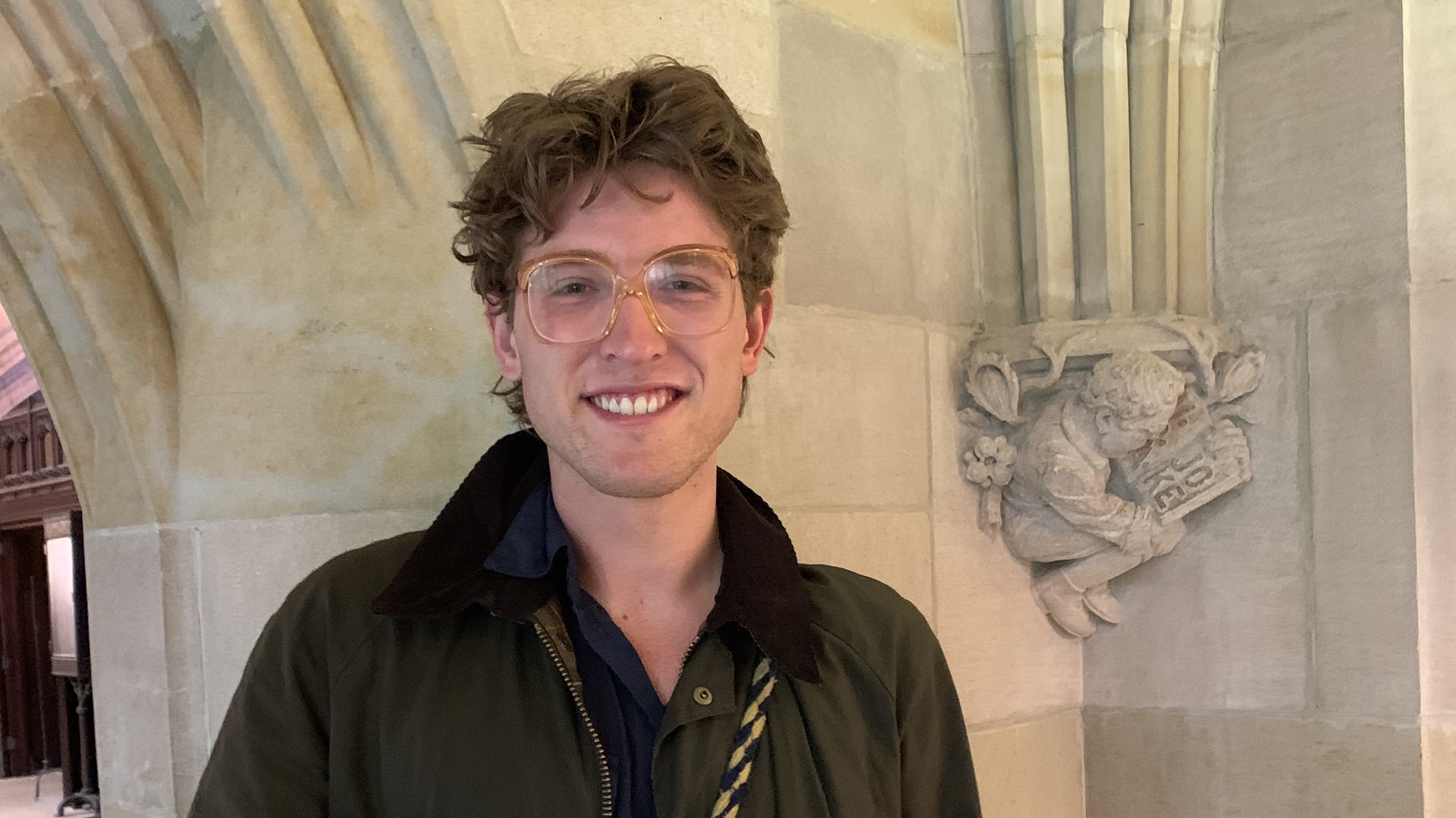 Young man with sandy brown hair and large amber-framed glasses, waring a dark shirt and jacket, smiles broadly and poses in front of carved column in the exhibition corridor of Sterling Memorial. The small carved figure is of a crouched man reading a book, one page of which has the carved word "Joke." 