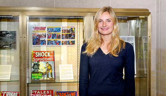 Female student with blond hair and navy dress standing in front of exhibit cases showing comic books and related research materials.