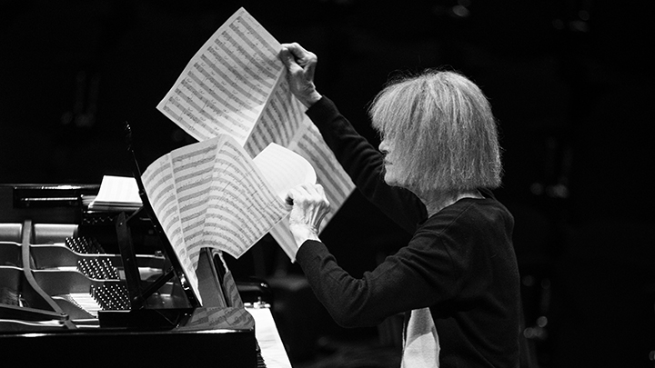 Woman with short white hair seated at piano lifting pages of music scores, one in each hand