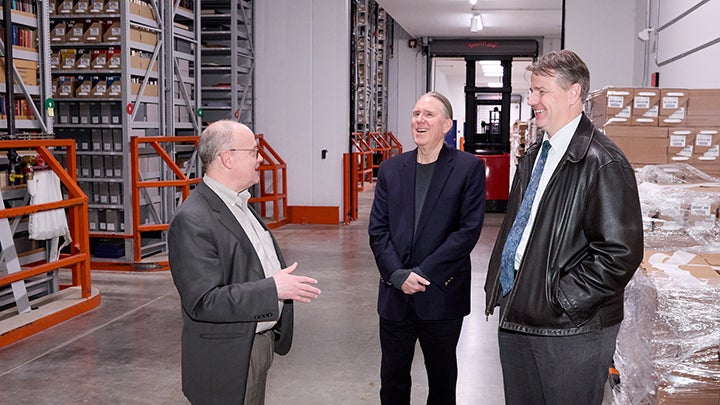 Three men talking in warehouse with tall grey shelving and orange bars. Short man is talking and two taller men are laughing.
