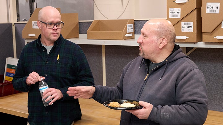 Bald man at left with dark glasses and black shirt holds plastic bottle listening to bald man at right who holds plate of food in left hand and gestures with right