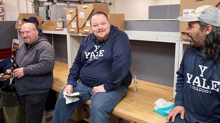 Two men sit on long wooden desk. Both wear blue shirts that read "Y Yale Bulldogs." Man at right wears cap. Third man at far left is bald stands and wears grey sweatshirt.