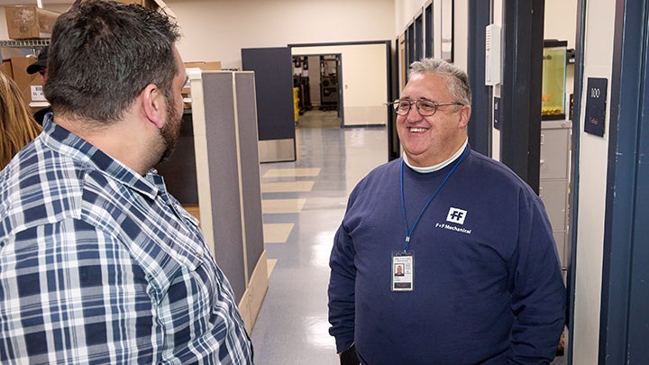 Two men talk. Man at left in black and white plaid shirt has back to camera. Man at right has dark blue sweatshirt with "FF" logo. He is smiling, has short, white hair and glasses. They are in long grey hallway.