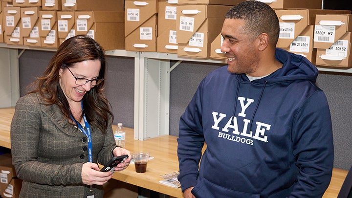 Woman with long dark hair and glasses leans over cell phone smiling. Man in blue sweatshirt looks on smiling. Shirt has white lettering that reads "Y Yale Bulldogs"