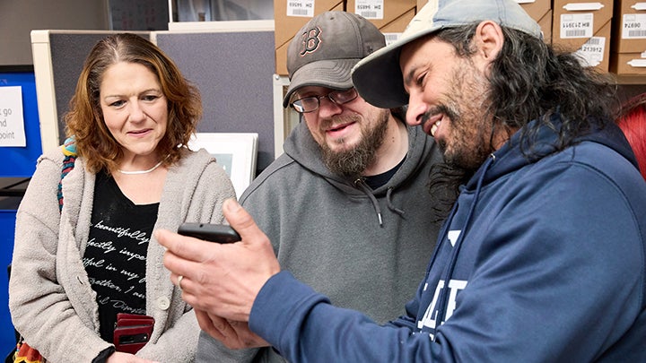 Woman at left and two men in sweatshirts and caps look at cell phone in hand of man at right