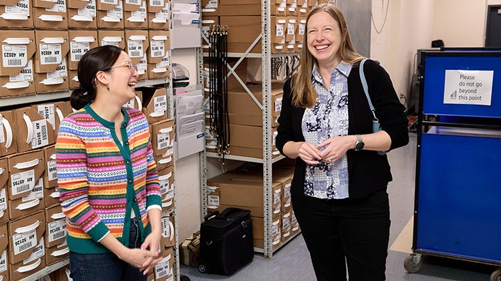 woman with black ponytail and multicolor striped sweater laughs with taller woman with long brown hair and black cardigan and pants. Behind them are shelves of brown boxes and a cart that reads "Please do not go beyond this point"