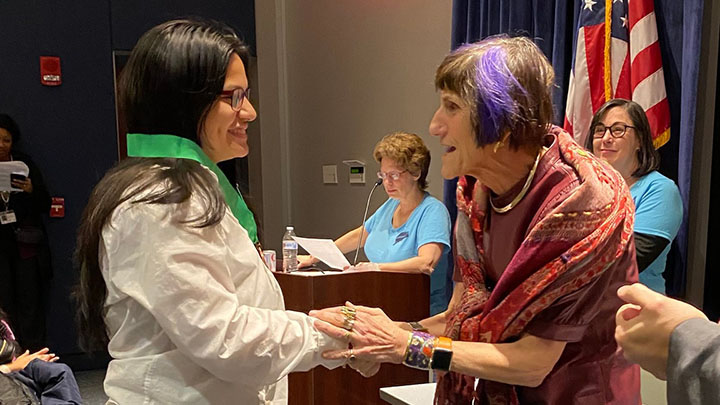 At left is young girl with long dark hair and glasses. Woman to her right, Rosa DeLauro, holds her hand. She has a purple streak in her short brown hair and wears a woven red shawl wrap around her shoulders. Two women with light blue shirts in background with American flag.