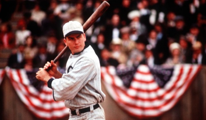 Baseball player with white pin-stripe hat and uniform holds bat over right shoulder looking into distance. Red, white, and blue bunting is draped behind him in front of the stands. The crowd looks on, although faces are blurred.