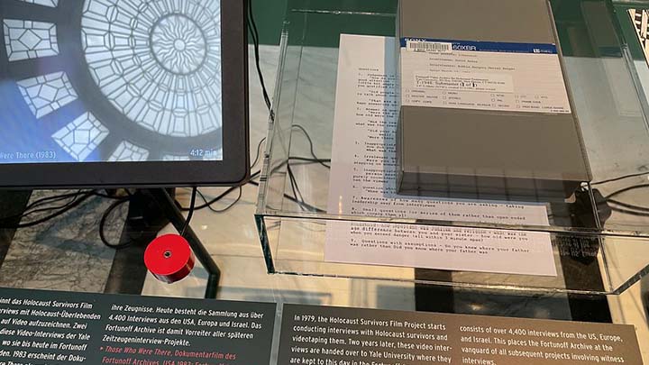 Closeup of exhibit display showing grey betacam cassette case on top of mylar surface; typed page shows through underneath; in upper left corner is a small video display showing a round decorative skylight. Descriptive text panels in German and English are visible along the bottom edge of the image.