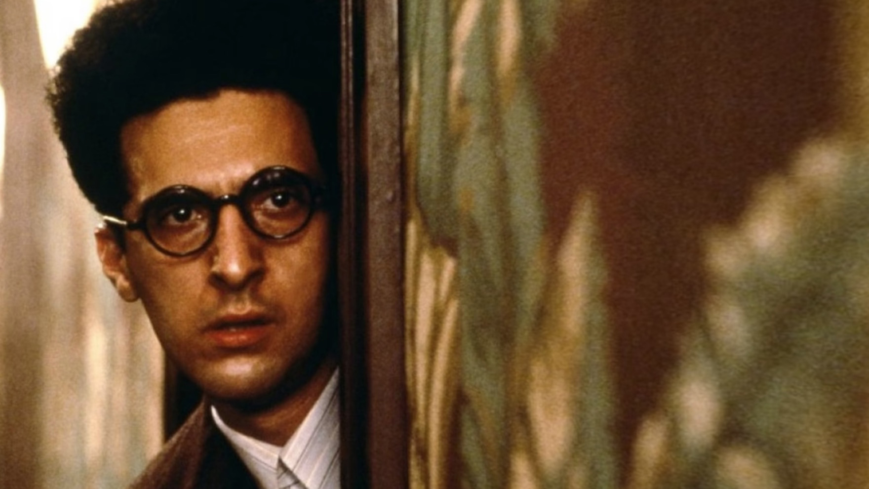 Film still showing head and shoulders of an anxious looking man with glasses and short curly hair  peering around the edge of a door