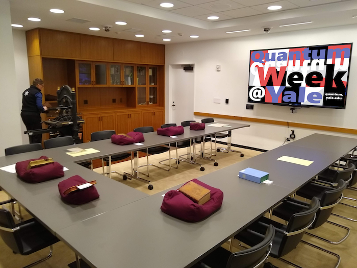Large U-shaped table in the Beinecke Library with several books on display, chairs around, and a large display on the wall that reads "Quantum Week at Yale"