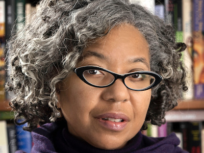 Author headshot of woman with curly gray hair and glasses in front of book shelves.