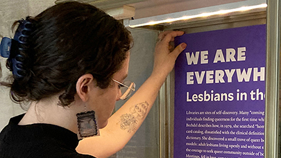 Curator Gabby Colangelo in dark shirt with hair tied back, mounting the introductory poster for her exhibit We Are Everywhere: Lesbians in the Archive