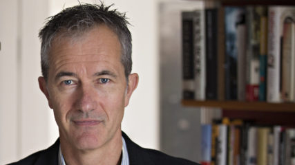 Portrait of Geoff Dyer in blue/white-striped button-down shirt and dark suit jacket, standing in front of wall of bookcases
