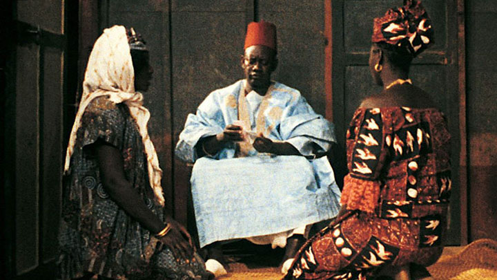 Two women in African dress and headdresses kneel on either side of man in white robe and red cap