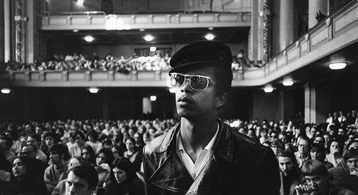 black and white image of an African American man wearing glasses, standing in front of a crowded room