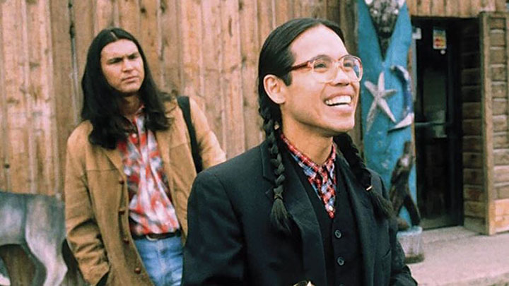 Two Native American men, one with long hair, one with braids and glasses smiling, stand outside rustic building.