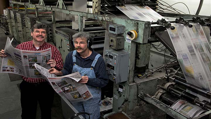 color photo of two men standing in front of a newspaper printing press holding newspapers