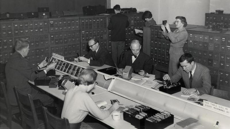 Black and white photograph of five faculty members reviewing photographic slides at a slide table with another woman standing in the background reviewing a slide and a man and woman standing in front of bank of file drawers, circa 1959.