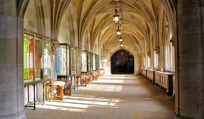 Sunlit stone hallway with arched ceiling and exhibit cases facing a wall of windows.