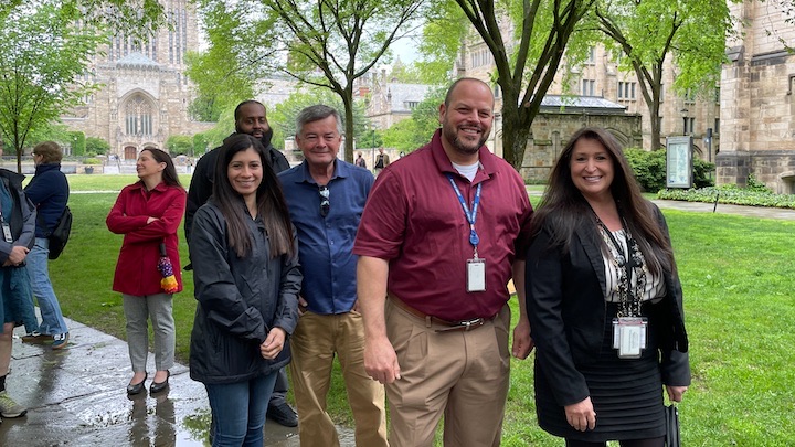 Seven staff waiting in line at the staff picnic with greenery and the stone facade of Sterling Library in the background. Five people are looking toward the camera. Sky is cloudy, pavement is wet, and several people are wearing raincoats or carrying umbrellas. 
