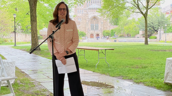 University Librarian Barbara Rockenbach  is standing in front of a mic on a stand holding a paper in her hands and speaking. She has shoulder length brown hair and glasses and is dressed in dark slacks and pink patterned  jacket background stands in front of a mic holding a sheet of paper in her hands. Next to and behind her can be seen  In the background can be seen  wet flagstone walk, green grass and trees, and the collegiate gothic facade in light colored limestone of Sterling Memorial Library.