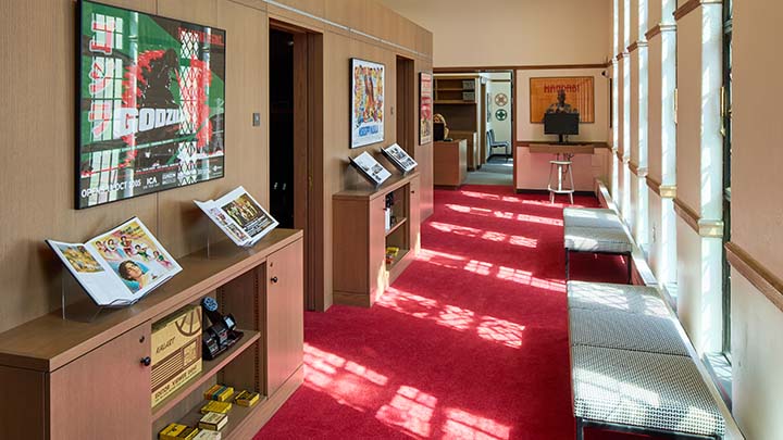 Red carpeted hallway with reflected light from side windows. Two bookcases on left display open books. A green, black, and red Godzilla film poster hangs on the wall. At right are a series of low cushioned stools.