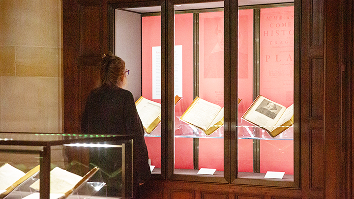 Woman in black sweater stands with back to camera, looking at a glass case displaying three open books. The book at far right shows image of William Shakespeare