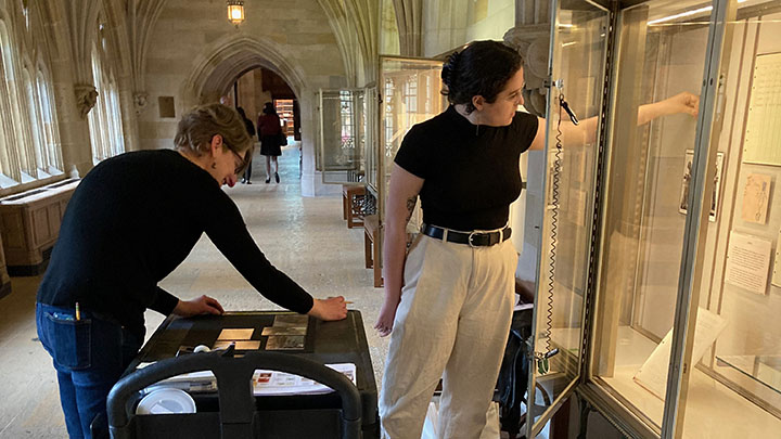 Two women working to install materials in glass cases that line the wall. The woman at left has short wavy brown hair and dark glasses and wears a dark sweater and jeans, bending over a work table and measure with a straightedge. The woman at right has dark hair pulled back in bun, wire glasses, black short-sleeve top, black belt and khaki pants. She reaches with one hand into the cases to post a paper document in the display.